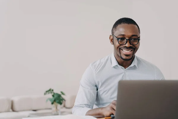 Smiling african american man software developer wearing glasses working on project online at laptop. Happy black guy programmer creating modern application or reading email with good news.