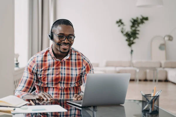Smiling african american young man wearing wireless headset with microphone, looking at laptop screen, learning online. Professional male teacher giving lesson to client, working remotely. E-learning.