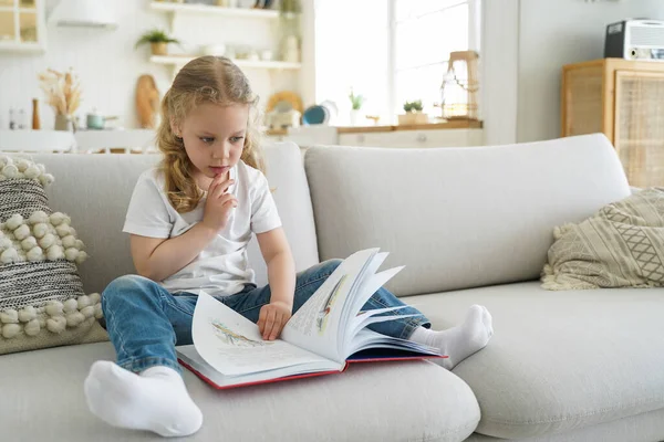 Cute pensive little child girl preschooler turns pages of fairytale book, sitting on sofa at home alone. Small kid daughter practicing reading in living room. Children's education concept.