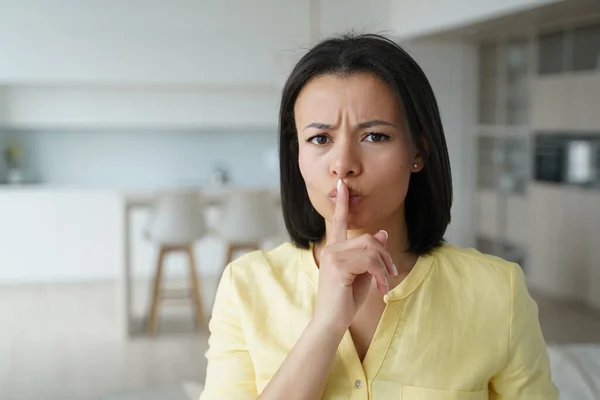 Frowning serious young woman showing hush gesture, standing at home, strict woman keeping finger on lips, demonstrating sign of silence, asking to keep privacy, secret private information.