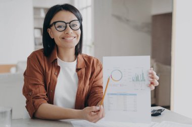 İş asistanı, istatistiksel veri grafiğine kalemle işaret ediyor. Gözlüklü, genç ve mutlu İspanyol menajer ev çalışma alanında çalışıyor. Çekici kız evrak işleriyle uğraşıyor ve gülümsüyor..