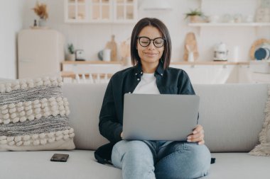 Gülümseyen, gözlüklü, dizüstü bilgisayarda çalışan, evdeki kanepede oturan serbest çalışan kadın. Genç bir kadın e-postaları, sosyal ağları kontrol ediyor, internetten alışveriş yapıyor. Uzak meslek, e-ticaret konsepti.