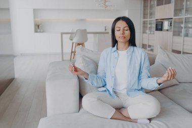 Sakin bir kadın yoga yapıyor, koltukta nilüfer çiçeği pozu veriyor. Serene genç kadın sakinliğini koruyor, evdeki kanepede meditasyon yapıyor, stresi azaltıyor. Meditasyon, sağlıklı yaşam tarzı konsepti.