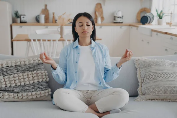 Calm woman practice yoga, sitting on sofa in lotus position, eyes closed, enjoy meditation at home. Female meditate, breathing for mental balance, relaxing. Healthy lifestyle, anxiety relief concept.