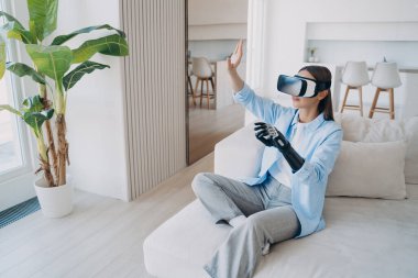Engelli kişi rehabilitasyona ve siber ortamda oynamaya gidiyor. Oturma odasında kanepede oturan beyaz bir kadın. VR gözlüklü engelli kız. Biyonik uzuv protezli ampüte.
