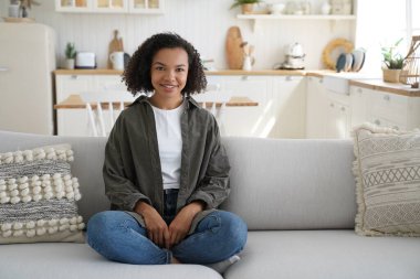 Rahat, genç, Afro-Amerikalı bir kız evinde Lotus pozisyonunda oturuyor, sağlıklı, gülümseyen bir kadın koltukta oturuyor, stressiz bir şekilde, rahat bir oturma odasında tembel bir hafta sonunun tadını çıkarıyor..