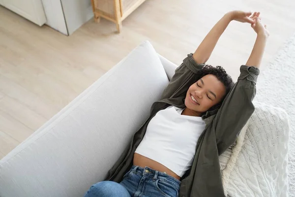 Happy pretty biracial girl resting lying on sofa at home with closed eyes, stretching body. Modern young female waking up after sleep, smiling, enjoying lazy weekend. Domestic life, wellness.
