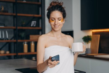 Sabah cilt bakımı sırasında akıllı telefon kullanan Latin kökenli genç bir kadın. Yüzünde göz bandı olan gülümseyen modern kadın evde telefondaki internet uygulamalarını kullanıyor..