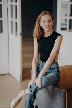 Kendinden emin, rahat kıyafetler giyen genç bir kadın evdeki kanepede oturuyor. Güzel model ince kız modern iç mekanda portre için rahat koltukta poz veriyor. Çevrimiçi mağaza tanıtımı.