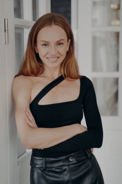 Sırıtkan, kendine güvenen, beyaz, çarpıcı, siyah bir üst giyen, kollarını kavuşturmuş, evinin kapısında duran bir kadın. Kameraya bakan sevimli bir kadının portresi..