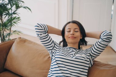 Huzurlu bir kadın rahat bir kanepede dinleniyor, elleri başının arkasında, sakin bir kadın dinleniyor, oturma odasında kanepede oturup temiz hava soluyor. Stres yok, sağlık konsepti.