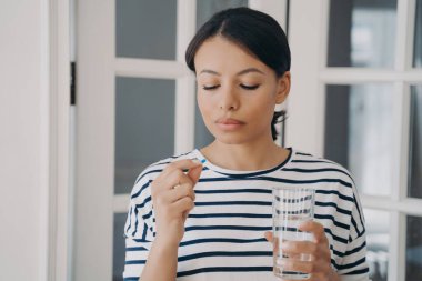 Sakin İspanyol kadın kapsülleri alıyor ve elinde bir bardak su tutuyor. Uyuşturucu alan sağlıksız bir kadın. Tıpla terapi. İlaçlar, vitaminler, farmakoloji. Ağrı kesici almak, hastalık küratörü olmak..
