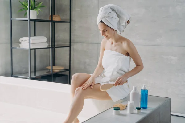 Pretty woman wrapped in bath towels massaging legs with wooden brush sitting on bathtub in bathroom. Female doing anti cellulite massage, scrub peeling body skin. Skincare, spa, self care concept.