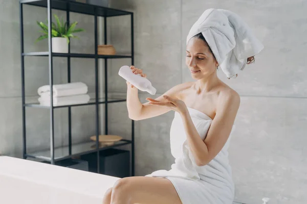 Hispanic woman using natural skincare oil in bathroom. Female wrapped in towel holding mockup bottle with moisturizing lotion sitting on bathtub. Body skin care treatment, organic cosmetics ad.