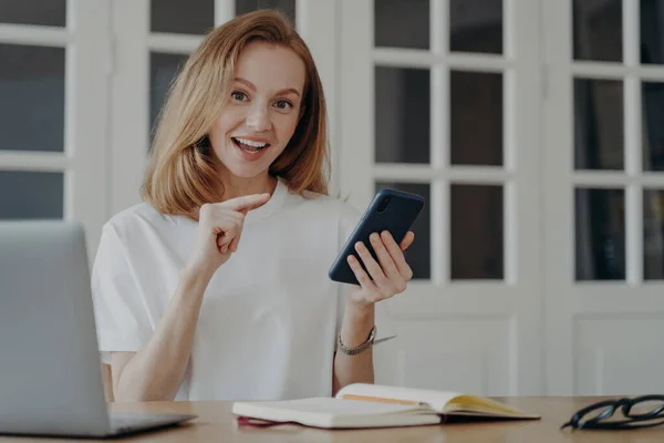 現代のスマートフォンを持った携帯電話で興奮した女性の指差指は ノートパソコンとオフィスの机に座っている 幸せな女性教育やビジネスのためのモバイルアプリを宣伝し 良いオンラインストアの提供 — ストック写真