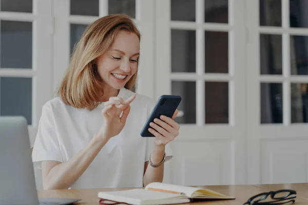 幸せなビジネスマンは 自宅のオフィスでスマートフォンを使用し 机に座っています 職場でビジネスやエンターテイメントのモバイルアプリを使用して 携帯電話で楽しんでいる女性を笑顔 インターネットを閲覧 — ストック写真