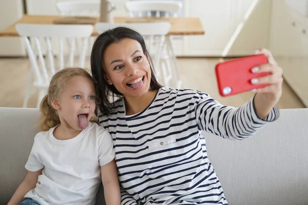 Playful mom and little child daughter holding phone, taking selfie photo, showing tongues together, recording funny video for social network. Small girl and mother having fun sitting on couch at home.