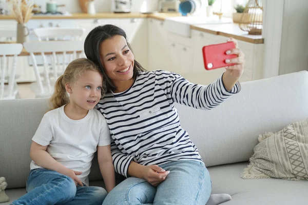 Happy mom and little daughter takes home selfie together, sitting on sofa, holding smartphone, looking at phone screen. Smiling mother and kid girl makes video call, chatting online, using mobile app.