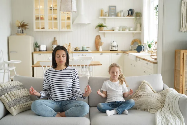 Calm mother with cute little foster daughter doing yoga exercises, sitting in lotus position on sofa together. Mom teaching adopted child girl to meditate at home. Healthy lifestyle concept.