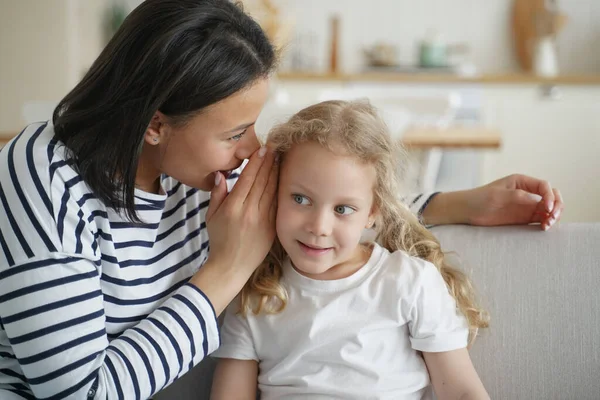 Female mother or babysitter telling secret, whispering to little cute kid daughter, sharing confidential information, gossiping. Mom and small child girl enjoying trustful conversation at home.