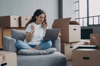 Düşünceli İspanyol kadın karton kutularla oturur taşınma gününde dizüstü bilgisayar kullanır, yeni ev için mobilya alır, tamir şirketini veya iç tasarım fikirlerini araştırır..