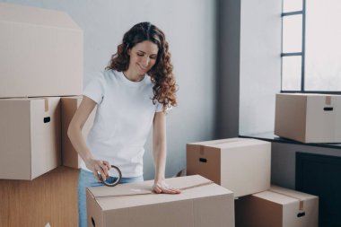 Gülümseyen genç İspanyol kadın yapışkan bant kullanıyor, karton kutu paketliyor. Taşıma işçisi kadın elinde koli bandıyla taşınma gününe ait eşyaları hazırlıyor. Teslimat, kaldırma konsepti.