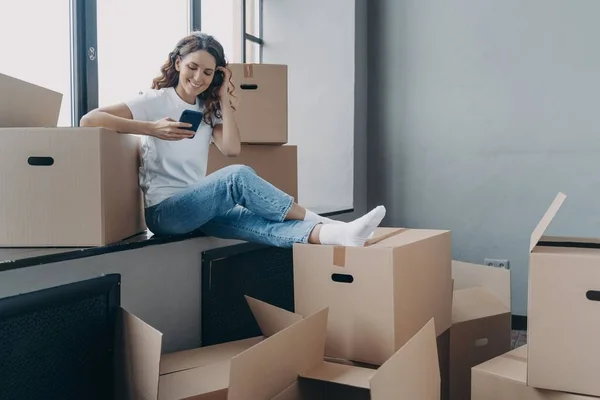 Smiling hispanic female sit on windowsill with carton boxes use smartphone apps buy modern furniture for first own new house or search moving company, arranging relocation. Ecommerce services, removal
