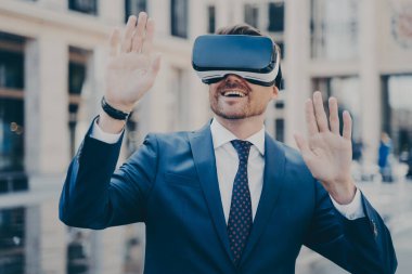 Ara verdiğinde takım elbiseli bir ofis çalışanı sanal gerçekliği VR gözlükleriyle keşfediyor, elleri havada dışarıda oturuyor, arka planda insanlar ve binalar bulanık, iş adamı oyun deneyiminin tadını çıkarıyor.