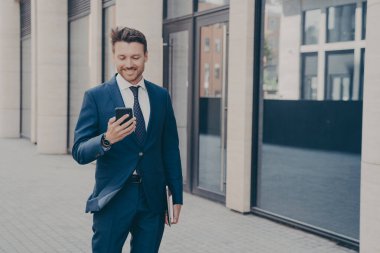 Kendinden emin genç erkek iş danışmanı modern akıllı telefonu tutuyor ve işten sonra dışarıda durup gülümsüyor, üniversiteden gelen mesajları okuyor ve projeyle ilgili iyi haberler veriyor.