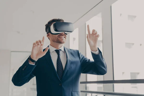 Excited office worker in vr headset or virtual reality goggles gesturing with hands, using innovative technologies for business at work, dressed in suit, smiling and touching objects in digital world