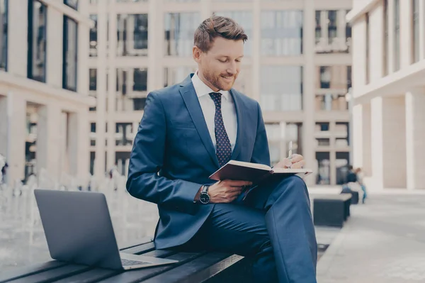 Relaxed and confident office worker in formal blue suit working outside while sitting on bench, using laptop and writing notes in agenda, city buildings in blurred background