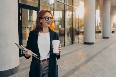 Resmi giyinmiş, doğal kızıl saçlı bir model, dokunmatik defter ve kahve tutuyor. Güneşli bir günde web sitesi paylaşımı için içerik yayınını düşünüyor.