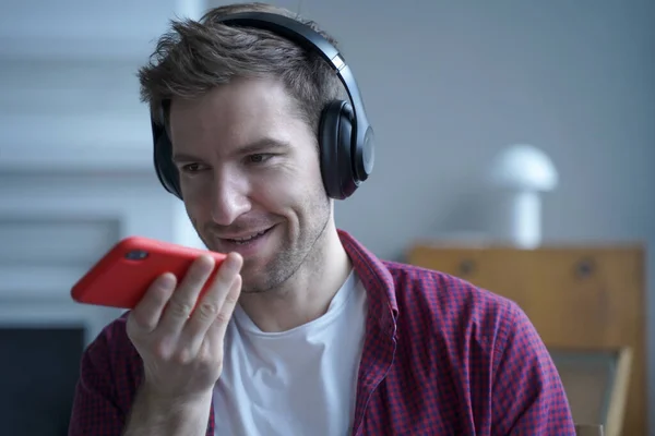 Young smiling german guy in headphones using translator app while studying online, holding mobile phone and talking on speakerphone with friend, recording voice message while spending time at home