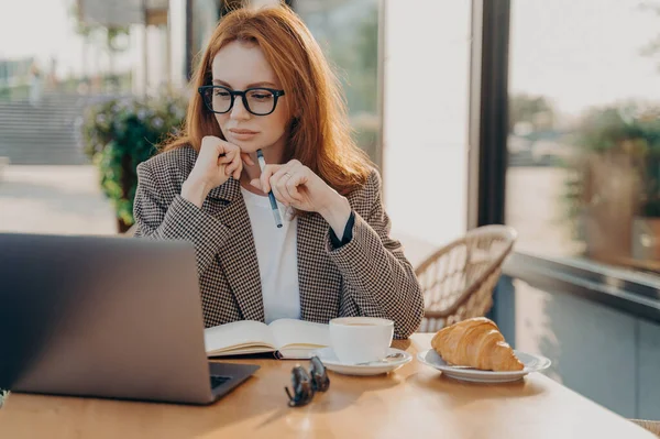 Serious ginger businesswoman in formal wear works in cafeteria develops new startup project poses against coffee shop interior makes notes in notepad works remotely. People technology freelancing