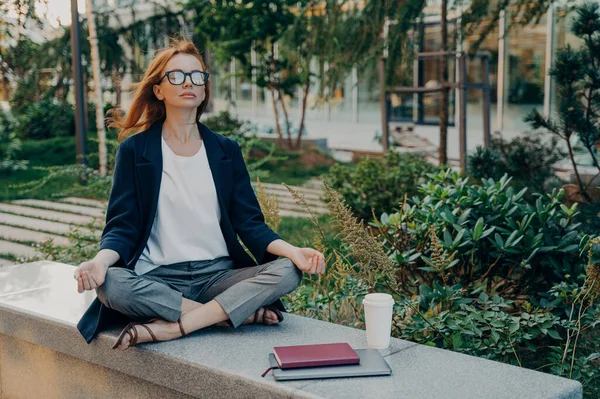 Peaceful redhead business woman dressed formally holding hands in mudra gesture, sitting in lotus pose on concrete bench and meditating with closed eyes, relaxing after remote work on laptop in park
