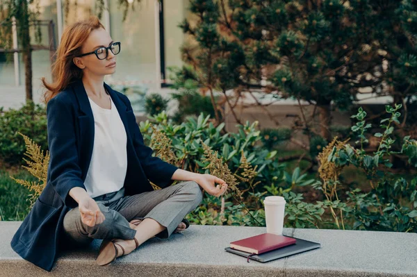 No stress. Calm red-haired woman with closed eyes relaxing while working remotely on laptop outdoors, mindful young business woman meditating with closed eyes, doing breathing yoga exercises outside