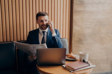 Mavi takım elbiseli memnun iş sahibi elinde gazeteyle telefonda konuşuyor, son haberleri ortağıyla paylaşıyor ve laptopa bakıyor, kafede çalışırken gelişmeleri bildiriyor.