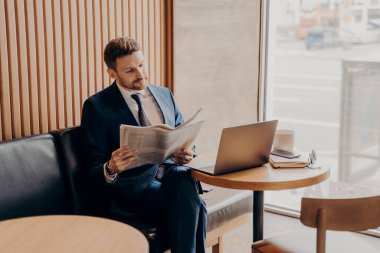 Mavi takım elbiseli ve beyaz gömlekli zengin genç işadamı, yerel gazetede şirketinin kuruluşuyla ilgili haberleri okurken önünde dizüstü bilgisayarla rahat bir kafede oturuyor.