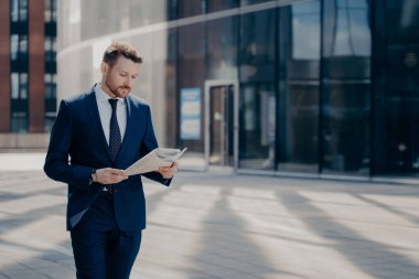 Resmi kıyafetli sakallı genç bir iş adamı elinde gazeteyle tek başına yürüyor, iş yerine giderken arka planda ofis binalarıyla son haberleri okuyor.