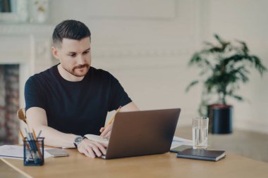 Ciddi sakallı adam, iş yerindeki iş yerindeki ofis içi pozlarını uzaktan giyip dizüstü bilgisayarda çalışan Webinar 'ı izlerken bilgilendirici notlar yazıyor. Modern çalışma alanı