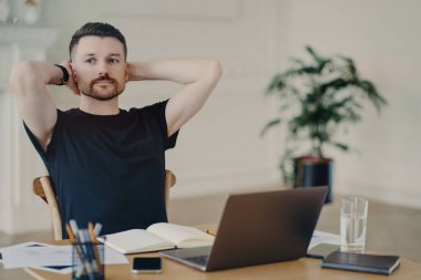 Düşünceli erkek serbest çalışıyor ellerini arkada tutuyor dizüstü bilgisayar başında poz veriyor düşünceli bir ifadeyle gelecek planları düşünüyor siyah t-shirt giyiyor..