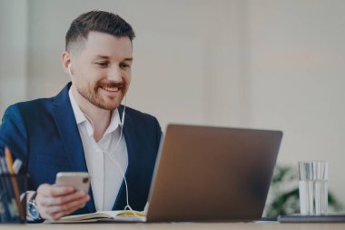 Mutlu erkek ofis çalışanı modern teknolojileri kullanıyor. Cep telefonu kullanıyor. Bilgisayarla video görüşmesi yapıyor, kulaklık takıyor ve resmi olarak giyiniyor.