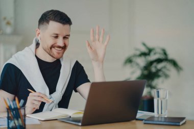 Neşeli sakallı adamın video konferansı var. Bilgisayarda avuç içi bilgisayar var. İş yerinde kablosuz kulaklık kullanıyor.