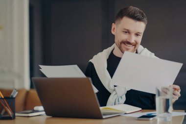 Serbest çalışan mutlu bir adam ya da sıradan giyinen bir iş adamı masada oturmuş, tatmin olmuş bir yüz ifadesiyle proje raporuna bakıyor. Dizüstü bilgisayarla rahat iş yerinde evrak işleriyle uğraşan erkek müdür
