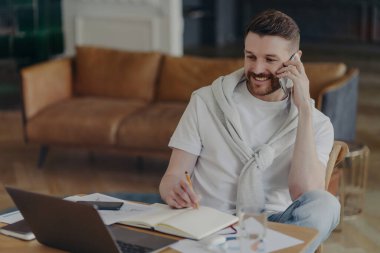 Neşeli serbest çalışan bir adam telefonla bilgi yazar not defterine not alır dizüstü bilgisayardaki iç dizüstü bilgisayar pozlarına dikkatle bakar. Teknoloji kullanıcısı