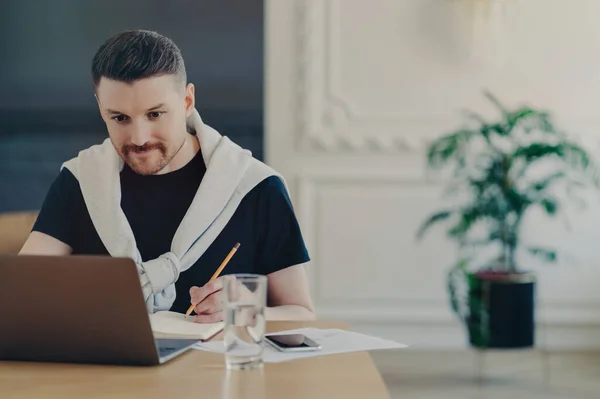 Young focused man freelancer in casual clothes looking at laptop screen, making some notes while sitting at his workplace at home, male manager gathering clients feedback or making project report