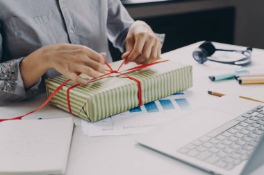 Genç iş kadınının paketleme ve kurdele Xmas hediyesiyle dekore ederken iş yerinde dizüstü bilgisayar ve sabit bir şekilde oturup Noel zamanı iş arkadaşlarına hediyeler hazırlarken görüntüsü.