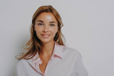 Güzel, çekici, mavi gözlü ve sağlıklı gömlekli bir kadının yakın plan portresi. Kameraya bakıyor, gülümsüyor, gri stüdyo arka planında soyutlanıyor.