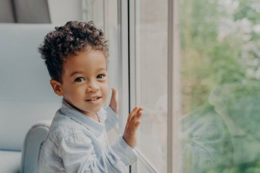 Melez kıvırcık saçlı açık mavi tişörtlü oğlan çocuğunun portresi. Pencere camına yaslanıp evde tek başına oynarken kameraya gülümsüyor.
