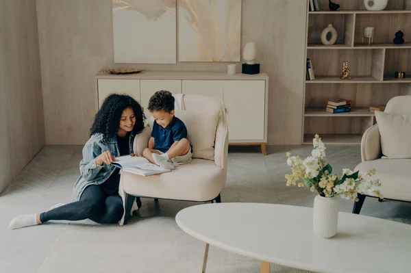 Little cute afro american kid sitting on chair with book next to his smiling and happy mother, learning english language together and having fun, happy mom and son spending quality time at home